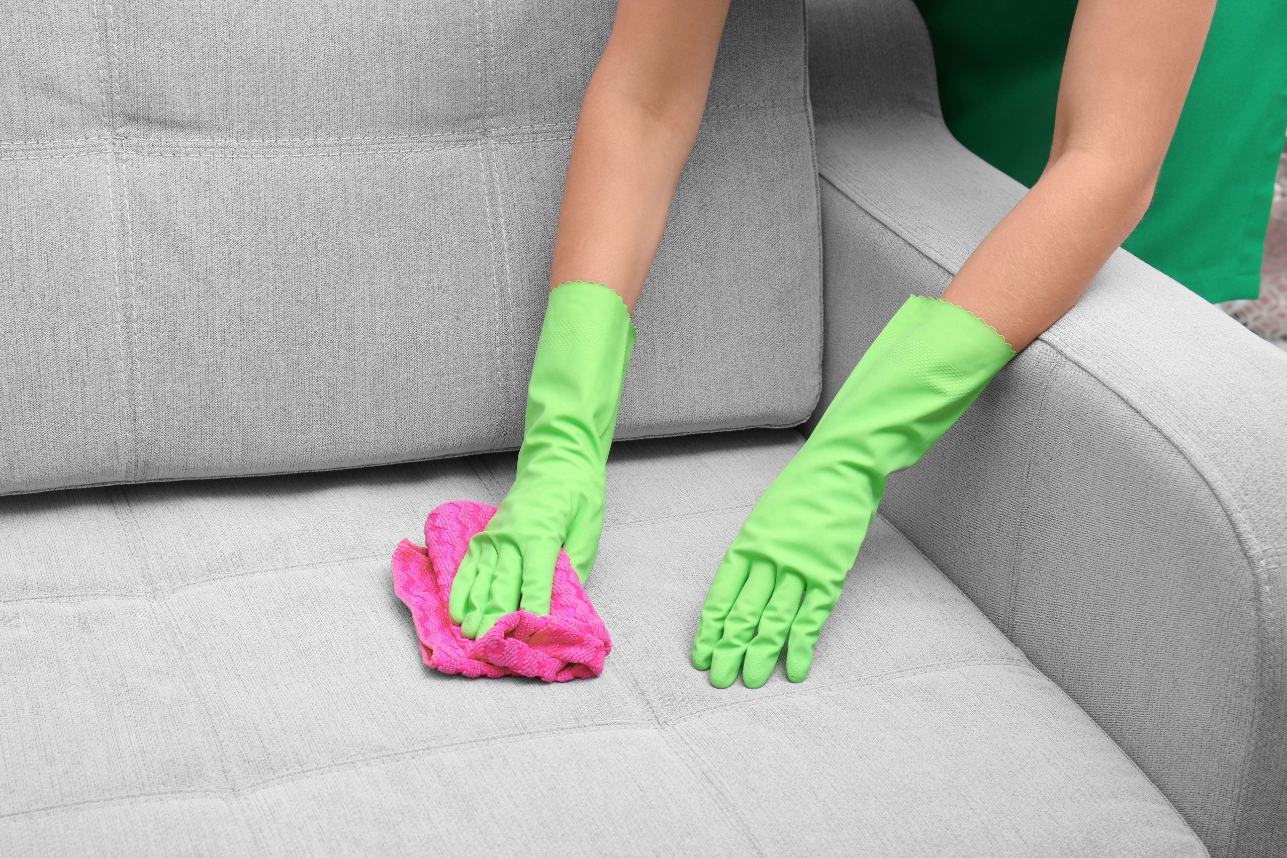 A imagem mostra a mão de uma pessoa com luvas limpando um sofá cinza.