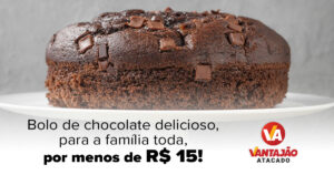 No centro da imagem, sobre uma forma branca elevada, está um bolo de chocolate redondo, com pedaços de chocolate no topo. O fundo da imagem é cinza.