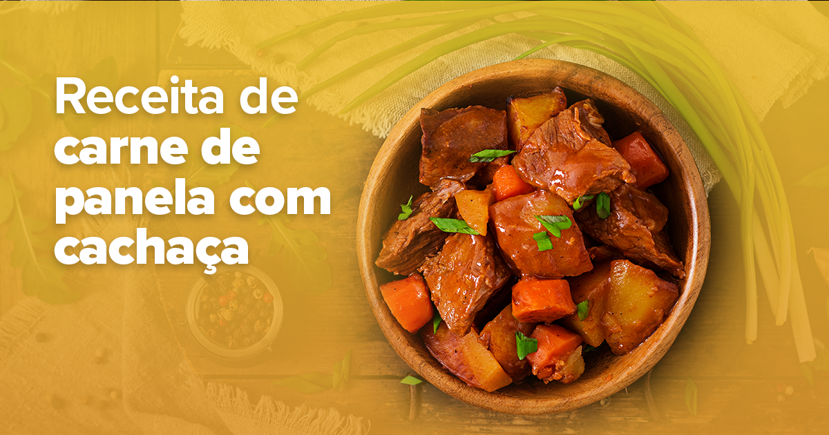 Dentro de uma tigela marrom arredondada, sobre uma mesa de madeira, estão pedaços de carne cozida com cenoura, cebola e salsinha. Sobre a mesa, ao redor da tigela, estão temperos como pimenta, cebolinha e alho.