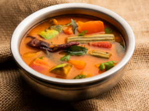 A imagem mostra uma cumbuca, sobre uma superfície com um pano marrom, com sopa dentro com uma coloração alaranjada. É possível ver pedaços de legumes diversos, como cenoura, e temperos verdes.