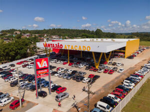 : Imagem aérea que mostra a fachada do mercado, com letreiro “Vantajão Atacado” em vermelho e amarelo, estacionamento em área aberta e redondezas da loja.