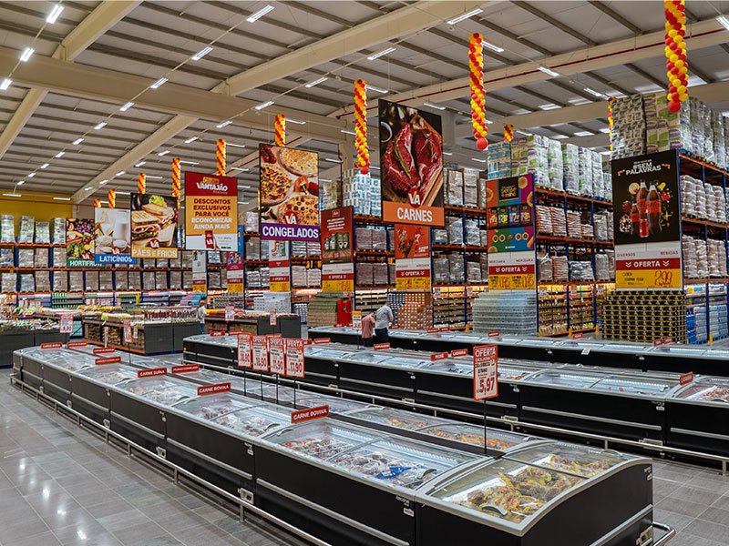 A imagem mostra o interior de um mercado, com diversos freezers e prateleiras altas lotadas com produtos, caixas e embalagens em geral. No teto, placas que indicam os produtos e balões vermelhos e amarelos.