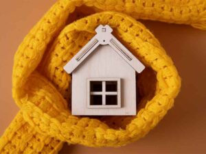 A imagem mostra uma miniatura de casa feita em madeira e pintada de branco. Ela está envolta em um cachecol amarelo.