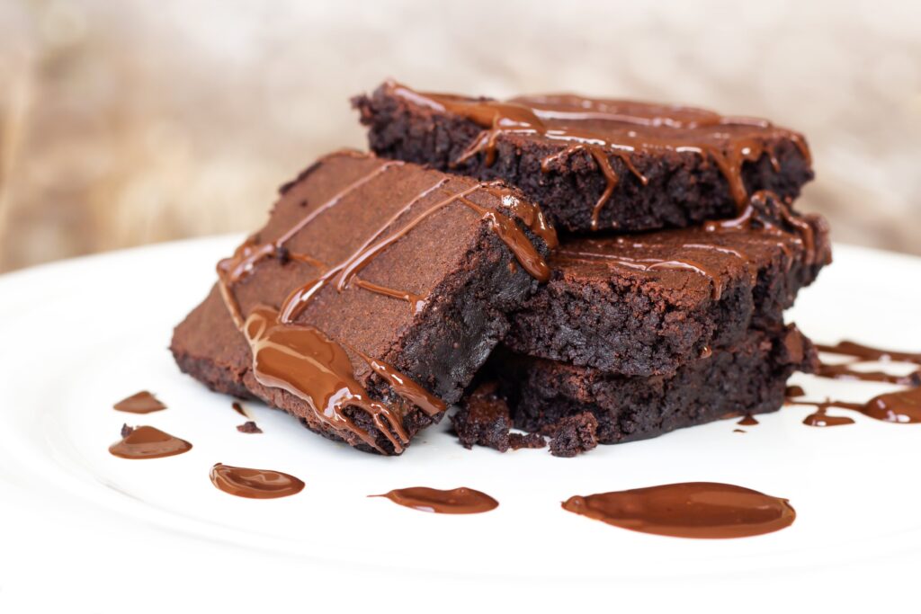 A imagem mostra quatro pedaços quadrados de brownie de chocolate com um pouco de chocolate derretido sobre eles. Eles estão sobre um prato branco. O fundo está desfocado e é marrom. 