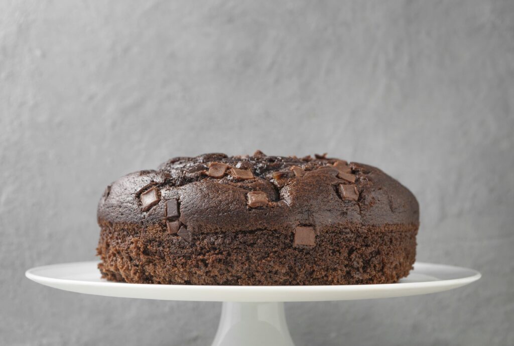 A imagem mostra um bolo de chocolate redondo e com pequenos pedaços de chocolate na parte superior. Ele está sobre um expositor branco. O fundo da imagem é uma parede cinza. 