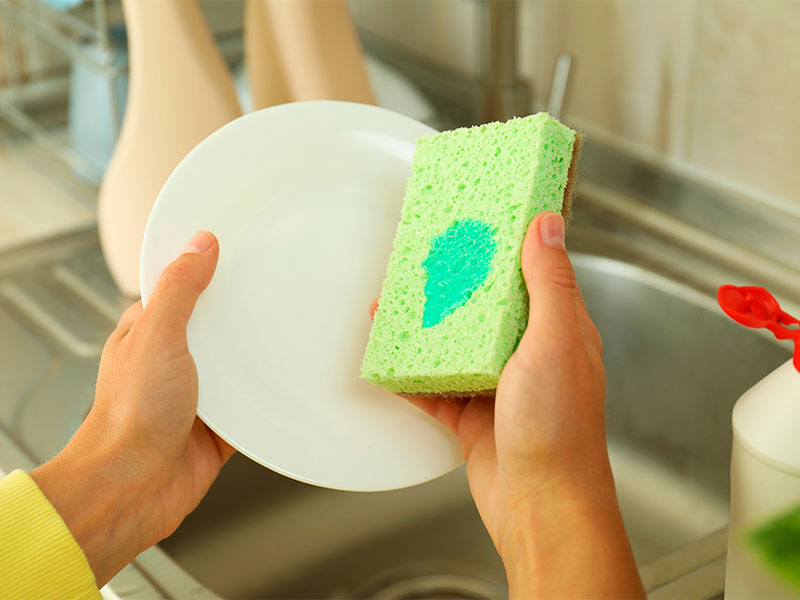 A imagem mostra duas mãos segurando um prato branco e uma esponja verde com detergente sobre ela. Ao fundo estão uma pia de inox e utensílios de cozinha. 
