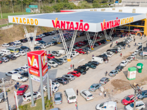 A imagem mostra uma vista aérea da nova loja do Vantajão Atacado. É um grande pavilhão nas cores amarela e branca, com letreiro grande à frente e vários carros no estacionamento frontal. É possível visualizar a cidade ao fundo.