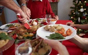 A imagem mostra uma mesa posta de Natal. Ao centro, está um prato com frango assado sendo cortado por um homem. Em primeiro plano, estão dois braços esticados segurando um prato com legumes. Ao fundo, nota-se uma árvore de Natal.