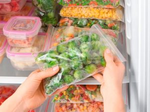 A imagem mostra as mãos de uma mulher segurando um pacote com legumes congelados. Ao fundo estão diversos alimentos congelados em potes e sacos plásticos.